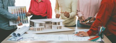 Profesyonel mimar takımının ev rengi seçimi hakkında beyin fırtınası yaparken, mimar da plan ve ev modelinin olduğu masadaki taslak belgeye yazı yazıyor. Değiştirilmiş.