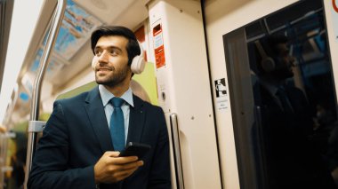 Akıllı yönetici trende dururken kulaklık takar ve müzik dinler. Profesyonel iş adamı, şarkıları dinlerken ve şarkı seçerken bölüme yaslanıyor. İş konsepti. Sevinçli..