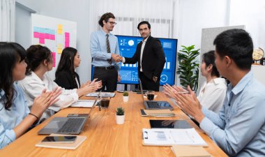 Toplantı odasında iş arkadaşıyla tokalaşan iki işadamı neşeli bir alkış veriyor. Analistlerden oluşan ekip, iş stratejik performansında kaydedilen önemli ilerlemeyi kutluyor. Habiliment