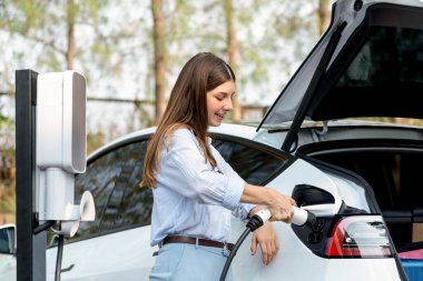 Genç bir kadın sonbahar gezisi sırasında sonbahar ormanı EV arabasında elektrikli araba için pilini şarj ediyor. Sonbahar tatilinde elektrikli araçla çevre dostu bir seyahat. Yükselt