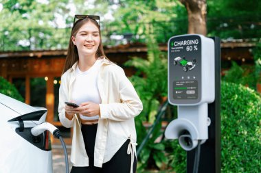 İlkbahar bahçesindeki açık kafede çalışan EV elektrikli araba şarjı, yeşil şehir sürdürülebilirliği ve çevre dostu EV otomobili için genç bir kadınla sürdürülebilir şehir seyahati elektrik ödüyor. Yararlı