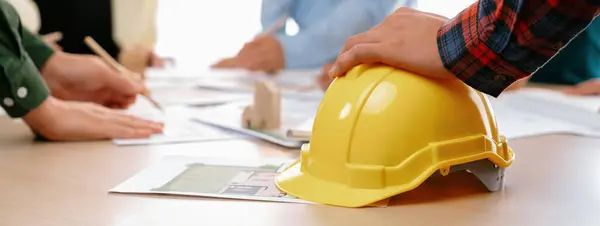 Professional architect team and engineer discuss about architectural project on meeting table with safety helmet, wooden block and architectural document scatter around. Closeup. Delineation.