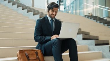 Kendine güveni tam bir iş adamı başarılı iş fikrini müzik dinlerken kutluyor. Happy yönetici projesi kulaklık ve takım elbise giyerken dizüstü bilgisayarla çalışıyor. Yatırımcı merdivenlerde oturuyor. Sevinçli..