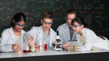 Çok kültürlü bir öğrenci mikroskop altında incelerken güzel bir kız da numuneyi inceliyor. BTMM sınıfında karatahta bilim teorisi deneyi yapan liseli çocuklara bakan öğretmen. Öğretim