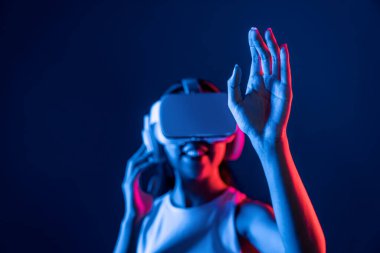 Çevresi siberpunk neon ışıklarıyla çevrili zeki bir kadın metaevrene bağlı VR kulaklık takıyor, gelecekçi siber uzay topluluğu teknolojisi. Sanal objeye el ile dokunan kadın. Halüsinasyon.