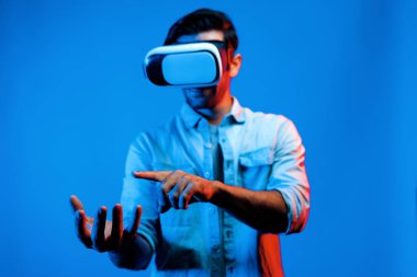 Sanal gerçeklik gözlüklü, sanal gerçeklikte hologram görüntüsü olan beyaz, zeki bir adam. Neon ışıklı arka planda gözlük ve kulaklık kullanarak bakıp işaret eden mutlu bir insan. Teknoloji. Sapma.