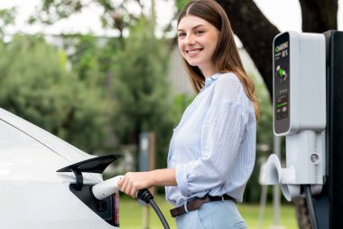 Açık yeşil şehir parkı manzaralı EV şarj istasyonundan genç kadın EV elektrikli araçları şarj ediyor. Çevre dostu şehir ulaşımı ve çevre dostu EV araba seyahati. Yükselt