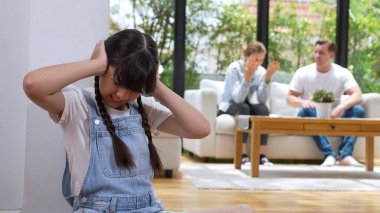 Stresli ve mutsuz genç kız köşeye kıvrılıp kulaklarını kapatarak arka planda ebeveyninin tartışmasını engelliyor. Evde aile içi şiddet ve travmatik çocukluk depresyona dönüşür. Eşzamanlama