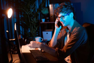 Genç iş adamı akıllı telefondan müşteriyi ya da iş arkadaşını arıyor, neon modern ofisteki yaratıcı sunum surround çalışma masasının bilgisayar ekranına bakıyor. Yeni proje kavramı. Toplayıcı.