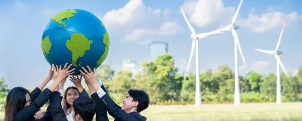 Earth day concept with big Earth globe held by asian business people team promoting environmental awareness using clean sustainable and renewable energy with wind turbine for greener future. Gyre