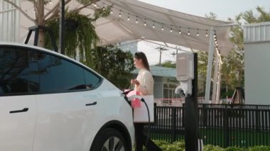 Genç kadın otoparktaki şarj istasyonundan EV araba aküsünü alışveriş çantasıyla şarj ediyor. Modern kadın, şehir içi seyahat tarzında çevre dostu elektrikli araçla alışverişe gider. Yararlı