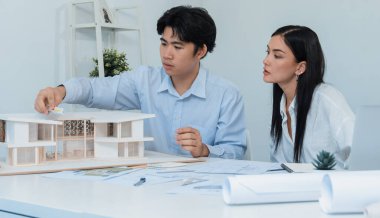 Professional male asian architect using ruler to measure house model length while young beautiful caucasian colleague using laptop to analyzed data on meeting table with house model. Immaculate. clipart