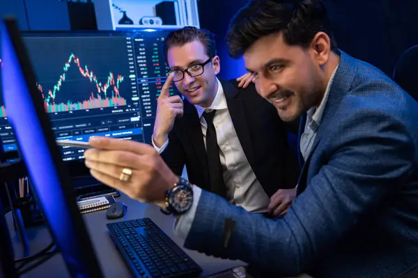 Stock image Two stock exchange traders discussing on dynamic successful investment graph, pointing trending of high profit in currency stock in financial data on monitor in neon light at workplace. Sellable.