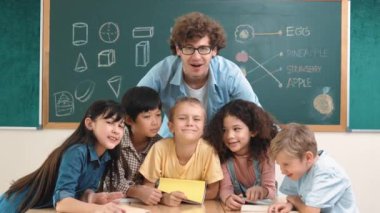 Kafkasyalı zeki öğretmen ve sınıftaki kameraya bakan çeşitli öğrenciler. Bir grup mutlu çocuk gülümserken eğitmen siyah tahtaya çıkıp cesaret vermek için sarılıyor. Eğitim. Pedagoji.
