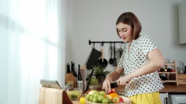 Kafkasyalı anne, modern mutfakta dizüstü bilgisayarda yemek tarifine bakarken salata yapmak için yemek hazırlıyor. Yetenekli ev bekçisi taze sebze kullanarak kahvaltı hazırlıyor. Pedagoji.