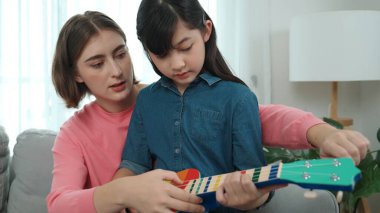 Kafkasyalı bir anne evde akustik müzik öğretirken ukulele çalan mutlu kız. Sevimli çocuk enstrüman öğreniyor. Kafkasyalı mutlu anne ve kız birlikte vakit geçiriyorlar. Pedagoji.
