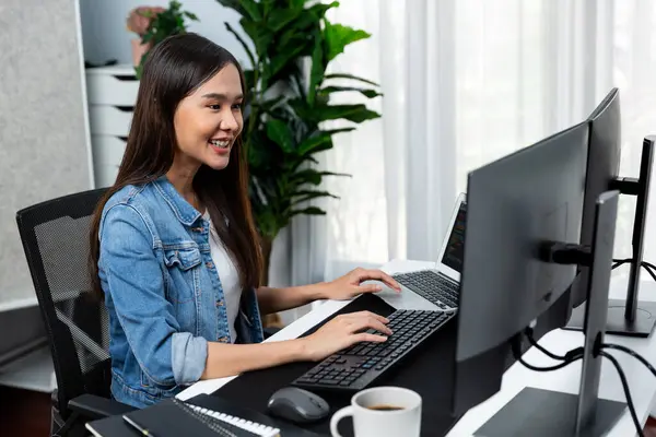 Smiling young Asian IT developer working software development information on pc screens and laptop, creating program coding for latest version application on website with firmware updated. Stratagem.