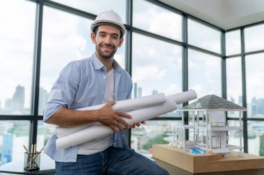 Modern ev projesini düşünen profesyonel mimar mühendisinin portresi güvenlik kaskı ve günlük kıyafetler giyerken. Akıllı iç mimar ev modelinin yanında proje planları tutuyor. İzleme.