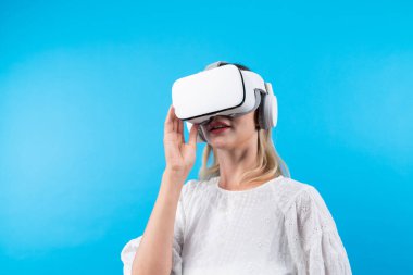 Mutlu kadın VR gözlüklerine bakıp mavi arka planda dururken şaşırdı ya da heyecanlandı. Beyaz pijamalı beyaz bir kız metaevrene bağlanmak için görsel gerçeklik gözlüğü kullanıyor. Kısıtlanma.