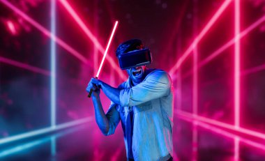 Lazer kılıcı tutan ve VR gözlük takan oyuncu. Beyaz adam, neon şatonun etrafında dikilirken görsel gerçeklik kulaklığı kullanıyor. Yenilik teknolojisi. Sapma.