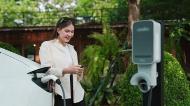 İlkbahar bahçesindeki açık kafede çalışan EV elektrikli araba şarjı, yeşil şehir sürdürülebilirliği ve çevre dostu EV otomobili için genç bir kadınla sürdürülebilir şehir seyahati elektrik ödüyor. Yararlı
