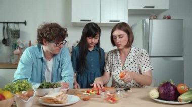 Çekici beyaz anne ve baba Asyalı kızıyla taze yemek pişirmek için birlikte vakit geçiriyorlar. Enerjik anne ve baba mutfakta salata yapmak için taze sebze hazırlıyor. Sağlıklı yiyecekler. Pedagoji.