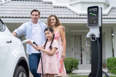 Mutlu küçük kız, ailesinin EV şarj istasyonundan elektrikli aracını şarj etmesine yardım ederken çevre dostu ve enerji sürdürülebilirliğini öğreniyor. EV araba ve modern aile kavramı. Eşzamanlama