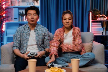 Mutlu çiftin oturduğu kanepe joysticklerle video oyunu oynuyor, meydan okuyan gülümseyen adam, evdeki modern neon ışıklı oturma odasında kazanmak için yumruğunu kaldırıyor. İnfobahn.