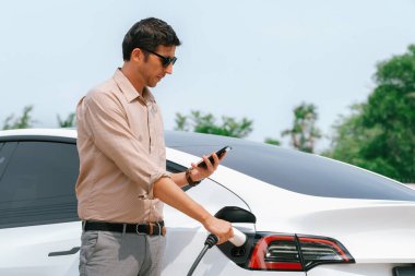 Genç adam EV otomobil şarj istasyonu Green City Park 'ta elektrik ödemek için akıllı telefonu kullanıyor. Modern çevre ve EV araçlı sürdürülebilir şehir yaşam tarzı. Yararlı