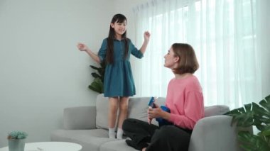 Annem gitar çalıp kıza bakarken yetenekli kız kanepede dans edip zıplıyor. Beyaz anne, çocuk oturma odasında dururken akustik müzik çalar. Aile eğlence konsepti. Pedagoji.