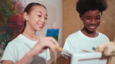 Yaratıcı sanat dersinde çeşitli çocuklar günlük tişörtleriyle tuvali boyuyor. Çok kültürlü mutlu lise öğrencisi renkli duvarın önünde dikilirken aynı tuvali çiziyor. Öğretim