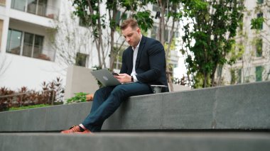 Yetenekli erkek lider Green City 'deki yatırım verilerini analiz ederken aynı zamanda dizüstü bilgisayarını kullanarak telefon gönderme raporu üzerinde çalışıyor. Parkta ya da şehir merkezinde otururken yatırımcı görünümlü mutlu pazarlama planı. Urbane.