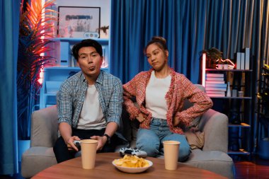 Mutlu çiftin oturduğu kanepe joysticklerle video oyunu oynuyor, meydan okuyan gülümseyen adam, evdeki modern neon ışıklı oturma odasında kazanmak için yumruğunu kaldırıyor. İnfobahn.