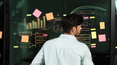 Profesyonel işadamı arkasını dönüyor ve kameraya bakıyor. Kollarını katlayıp duvara bakıyor. İstatistiksel ve yapışkan notlarla. Kameraya gülümserken kollarını kavuşturan beyaz erkek lider Tracery