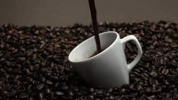 Fresh espresso was poured in white cup with pile of coffee bean. Pouring black coffee in the cup with brown background and seed scattering around on wooden table. Close up. Slow motion. Comestible.