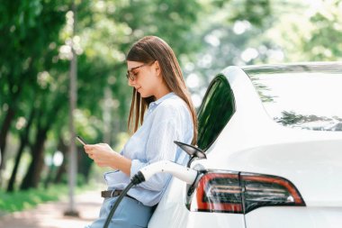 Ulusal Park veya Yaz Ormanı 'ndaki tatil gezisi sırasında EV şarj istasyonundan elektrik aküsü almak için akıllı telefon internet bankacılık uygulaması kullanan genç bir kadın. Yükselt