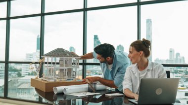 Akıllı mimar mühendis ev modelini incelerken meslektaşı da laptop analiz verilerini kullanıyor. Modern ofisteki ev modeli inşaatında profesyonel tasarımcı takımı birlikte çalışıyor. İzleme