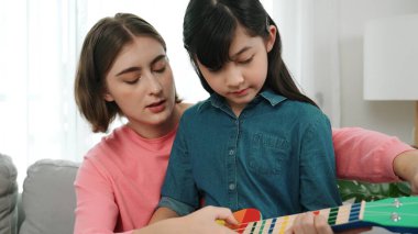 Kafkasyalı bir anne evde akustik müzik öğretirken ukulele çalan mutlu kız. Sevimli çocuk enstrüman öğreniyor. Kafkasyalı mutlu anne ve kız birlikte vakit geçiriyorlar. Pedagoji.
