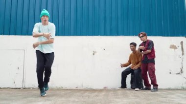 Genç sokak dansçısı hip hop tarzında dans ediyor ve çok kültürlü arkadaşları duvarın arkasında tezahürat yapıyor. Çekici yakışıklı adam mavi arka planda kollarını uzatıyor. Açık hava sporu 2024 hiphop..