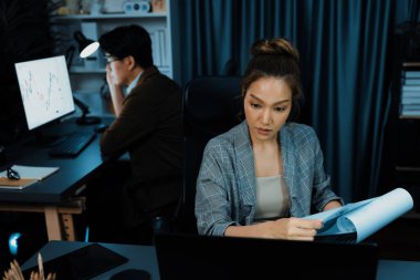 Stresli yüzü olan bir iş kadını evrak dosyasını incelerken, iş arkadaşı da gece geç saatlerde modern ofiste borsa alım satımı yapıyor ve şirket projesi üzerinde çalışıyor. İnfobahn.