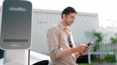 Ağır çekimde çalışan adam, alışveriş merkezinin otoparkındaki kamu EV şarj istasyonunda elektrik için akıllı telefon kullanıyor. EV aracıyla modern çevre ve sürdürülebilir kentsel yaşam tarzı