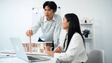 Profesyonel Asyalı erkek mimar ev modelinin uzunluğunu ölçmek için cetvel kullanırken, beyaz tenli genç meslektaşı ev modeliyle toplantı masasındaki verileri analiz etmek için dizüstü bilgisayar kullanıyor. Tertemiz..