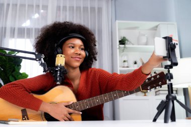 Akıllı telefon ayarlayan güzel Afrikalı kadın nüfuzlu sunucu kanalı yayında gitarla şarkı söylüyor. Canlı sosyal medyada müzik blogcusu zamanı. Ses yaratıcısı kavramı. Tatlandırıcı.