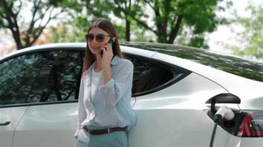 Doğal orman ya da milli parktaki araba yolculuğu sırasında elektrikli araba pili şarj ederken kadın telefonda konuşuyor. Tatil ve tatil boyunca çevre dostu seyahat. Yükselt