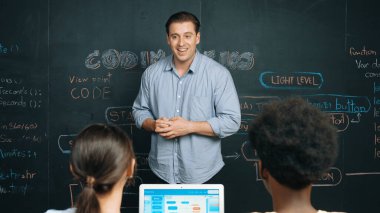 Tahtada çeşitli öğrencilere mühendislik kodları öğreten yakışıklı bir öğretmen. Mutlu çok kültürlü liseli çocuklar dizüstü bilgisayar kullanırken öğreniyor ya da programlama veya kodlama sistemini öğreniyor. Öğretim.