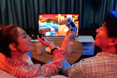 joystickli elleri çapraz oynayan bir çift arka tarafta TV ekranında dövüş oyunu oynuyor, modern rahat evinde neon ışıklı oturma odasında kanepesinde oturuyor. İnfobahn.