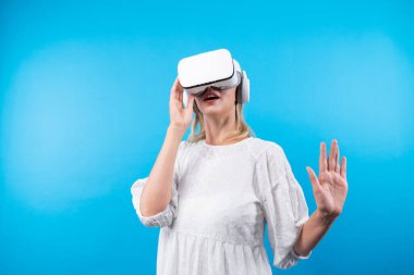 Mutlu kadın VR gözlüklerine bakıp mavi arka planda dururken şaşırdı ya da heyecanlandı. Beyaz pijamalı beyaz bir kız metaevrene bağlanmak için görsel gerçeklik gözlüğü kullanıyor. Kısıtlanma.