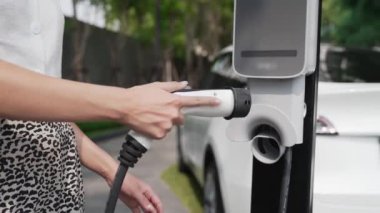 Şehir merkezindeki EV şarj istasyonunda elektrikli araba aküsünü yavaş çekimde şarj eden kadın elektrikli aracın iç organları için sürdürülebilir yeşil ve yenilenebilir temiz enerji yaşam tarzına sahip.