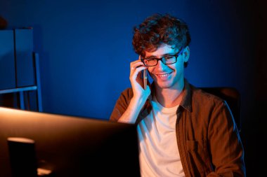 Genç iş adamı akıllı telefondan müşteriyi ya da iş arkadaşını arıyor, neon modern ofisteki yaratıcı sunum surround çalışma masasının bilgisayar ekranına bakıyor. Yeni proje kavramı. Toplayıcı.