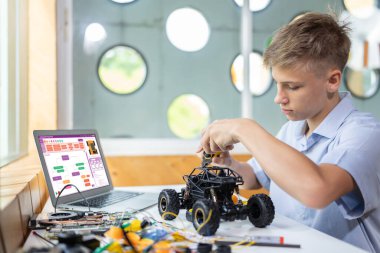Sarı saçlı çocuk BTMM sınıfında teknoloji öğrenirken kerpeten kullanarak robot araba tamir ediyor. Masaya dizüstü bilgisayar, kumanda, elektrik kablosu, şarj aleti ve robot araç koyun. Öğretim.